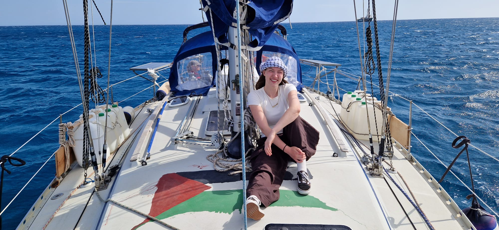 A person wearing a white T-shirt, brown trousers, and a keffiyeh-patterned hat sits on the deck of a sailboat at sea. The boat features a Palestinian flag painted on its stern, with ropes, jerry cans, and a blue canopy visible. The ocean stretches out under a clear sky in the background.