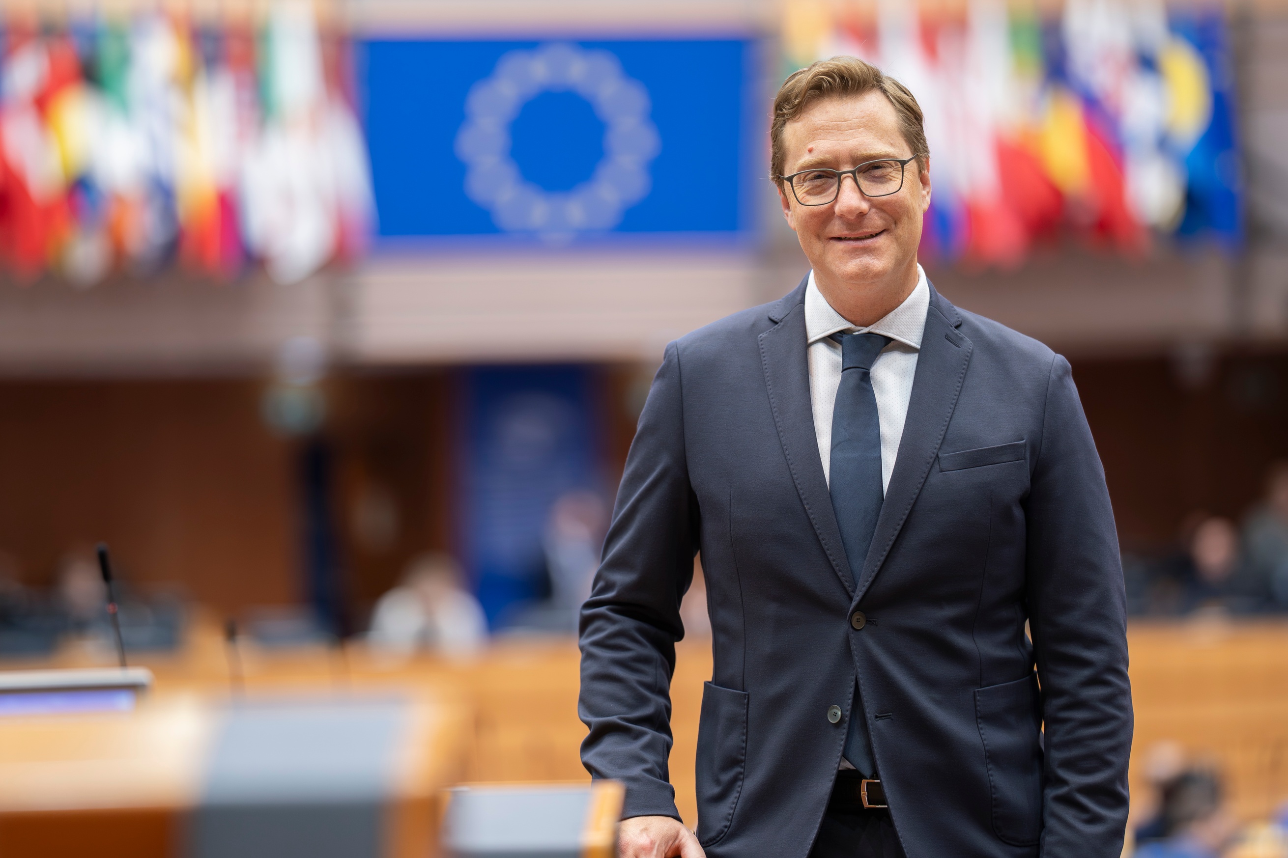 Vladimir Prebilič standing on the right of the photo, the hemicycle of the European Parliament blurred behind him with a flag of the european Union