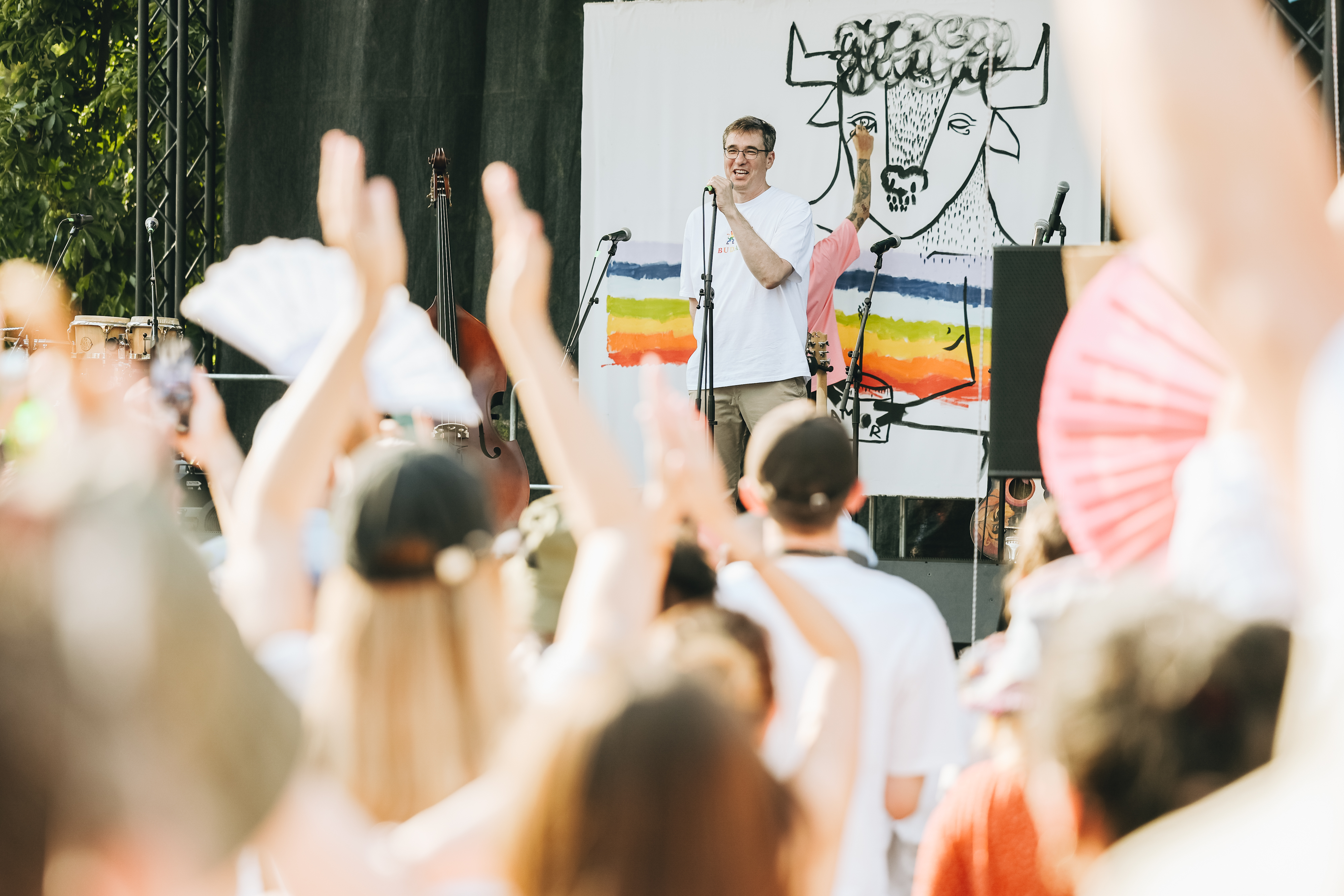 The mayor karacsony, white t-shirt, standing on a stage with a mic in front a crowd cheering on a sunny day. Behind him a man paints a mural.