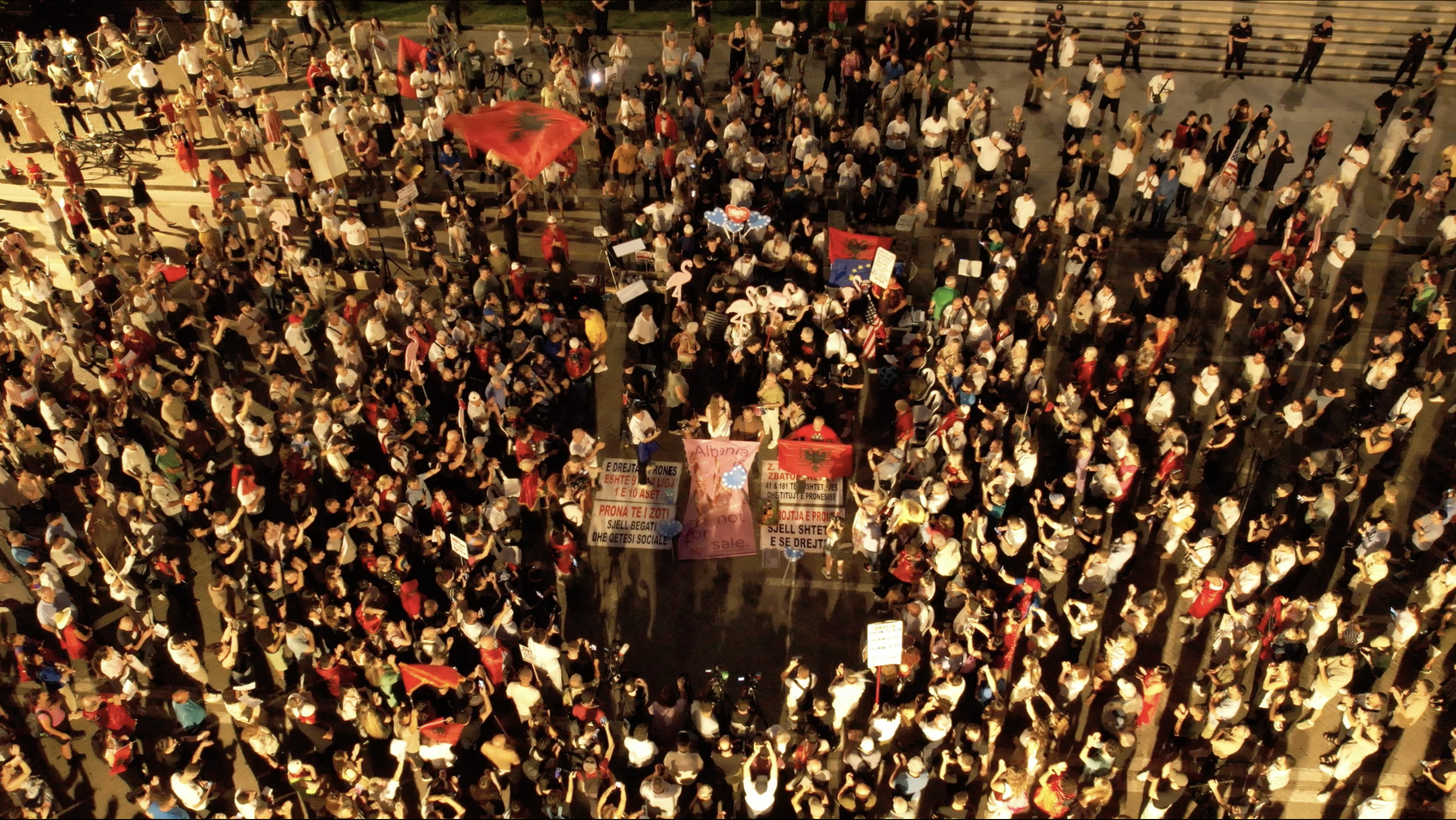 Crowd gathered at night, circling a small lectern for speeches, with albanian flags.