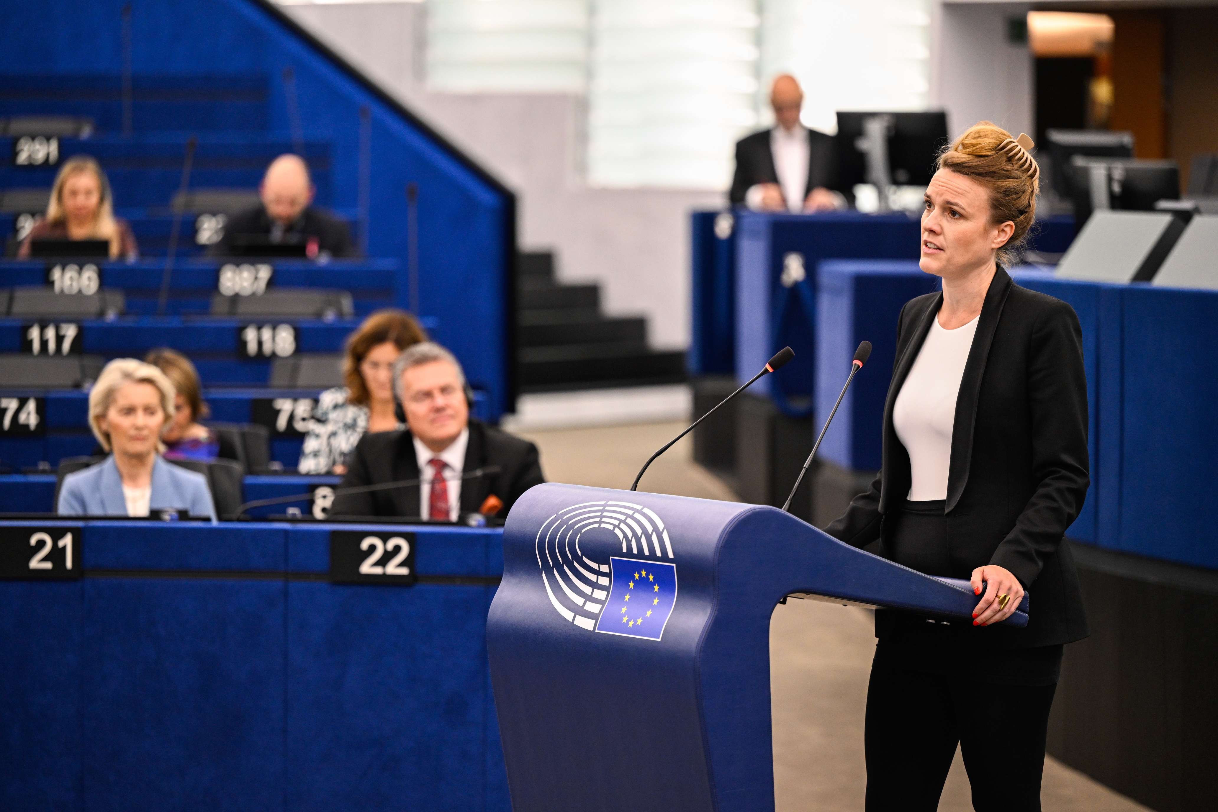Terry reintke standing in front of MEPs and Ursula Von der Leyen, speaking behind a lectern with the european commission logo