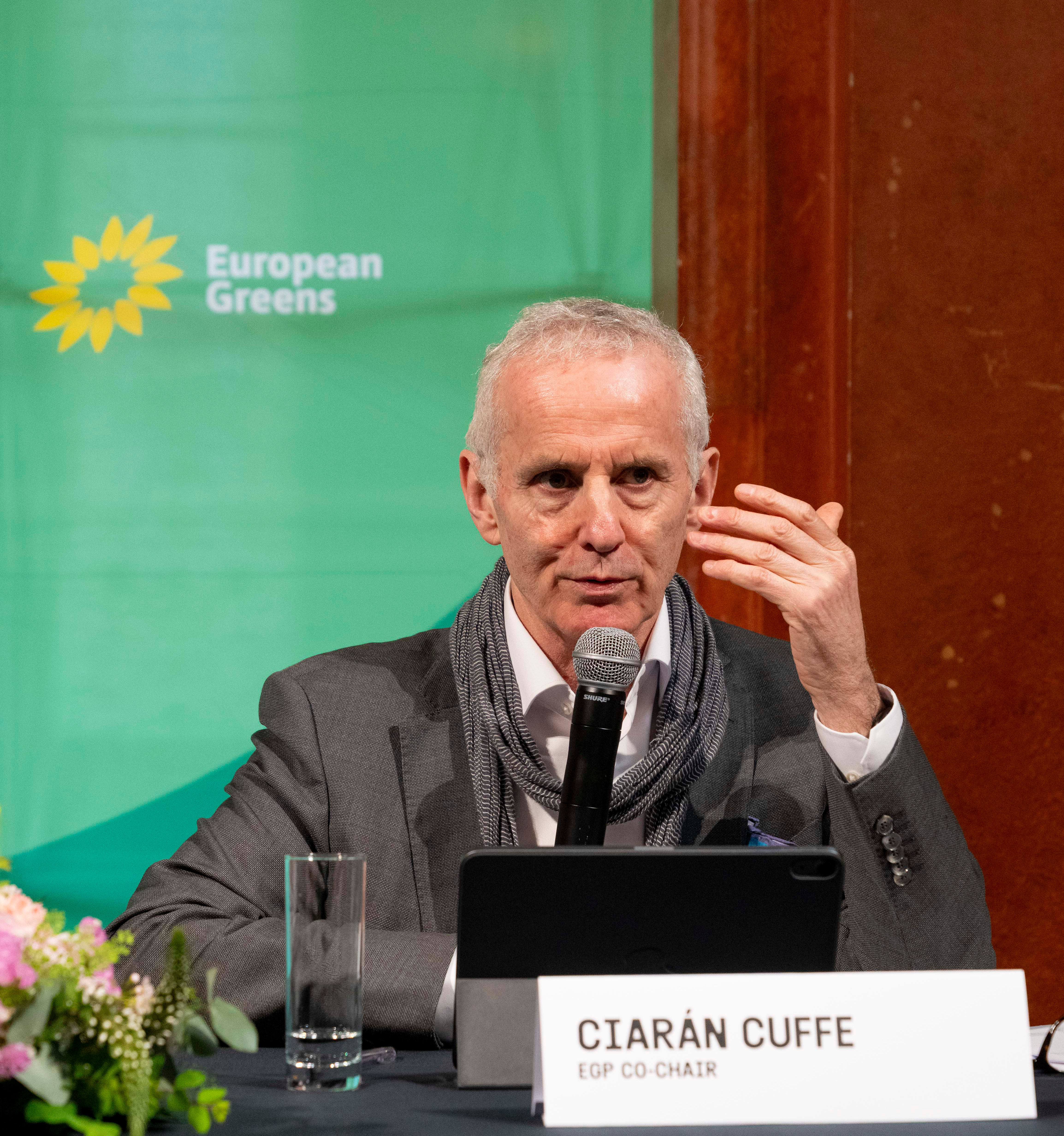 Ciarán Cuffe sitting at a presidium table in front of a european Greens backdrop with logo, with a name tag reading "Ciarán Cuffe Co chair" in front of him