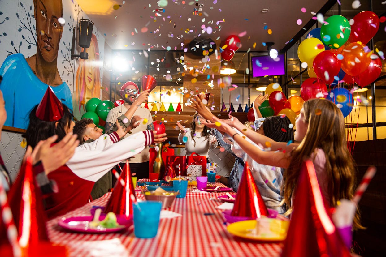 Group of kids in a Strike party room, throwing raining confetti in celebration. The party room is decorated with balloons and party supplies.