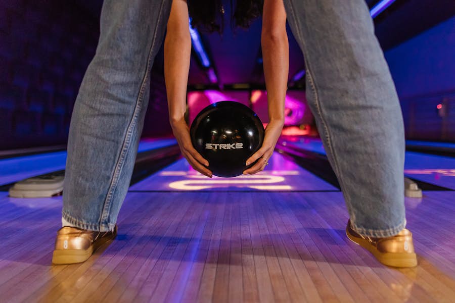 Person getting ready to bowl on the Strike lanes, tunnel ball style. They are holding a black bowling ball with the Strike logo on it, and wearing gold Strike bowling shoes.