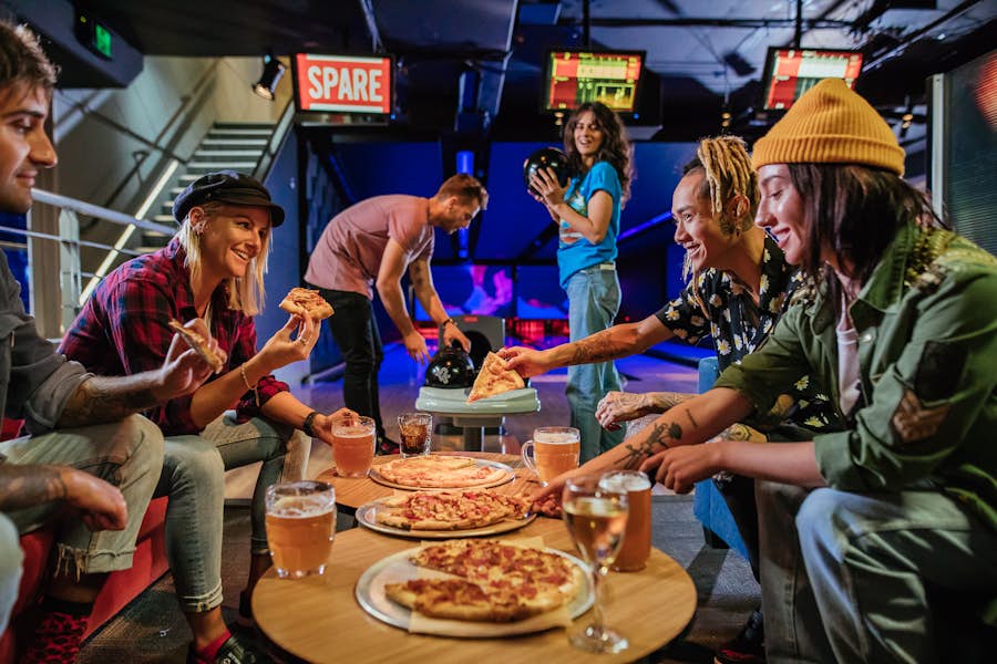 Group of friends eating and drinking while 2 people are bowling in the background