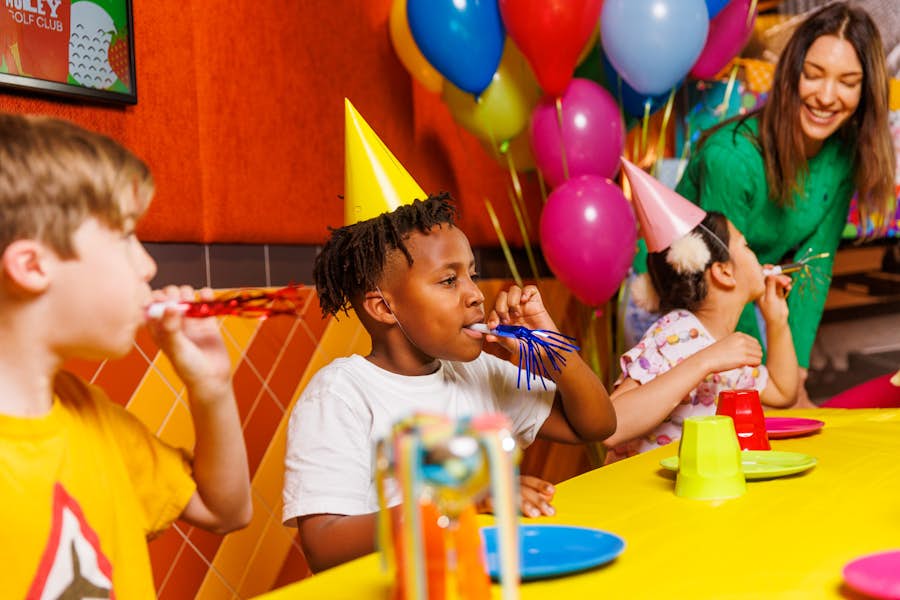Children wearing party hats in a Holey Moley Party Room, blowing into party noise makers.