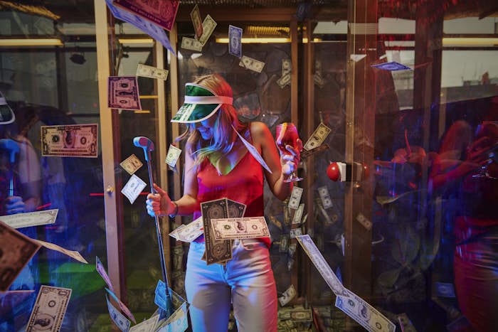 Woman wearing a green Holey Moley visor and holding a putter in one hand and a cocktail in the other smiling while fake money surrounds her in the 'Making It Rain' hole at Holey Moley.