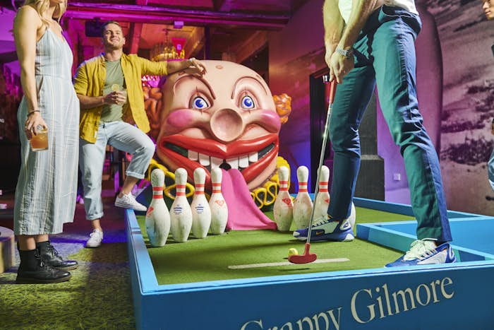One person is lining up to putt into a giant clown's face on the Crappy Gilmore mini golf hole, while two friends watch on.