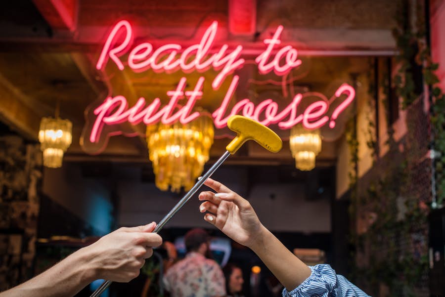 Close up of one person's hand passing a putter to another person. There is a neon sign in the background that reads "Ready to putt loose?.