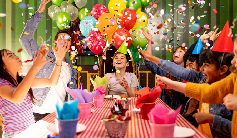 Group of kids/adults in an Archie Brothers party room, throwing raining confetti in celebration. The party room is decorated with balloons and party supplies.