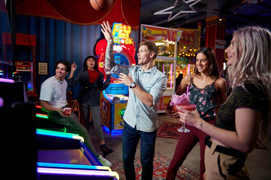 A group of 5 people, with one of them shooting a basketball on the hoops game, while the four others watch on with drinks in hand.