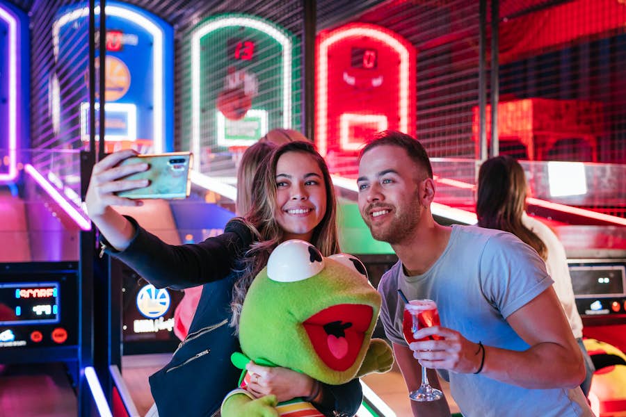 Two people taking a selfie at Archie Brothers, while one is holding a plushie of kermit the frog from the redemption store.