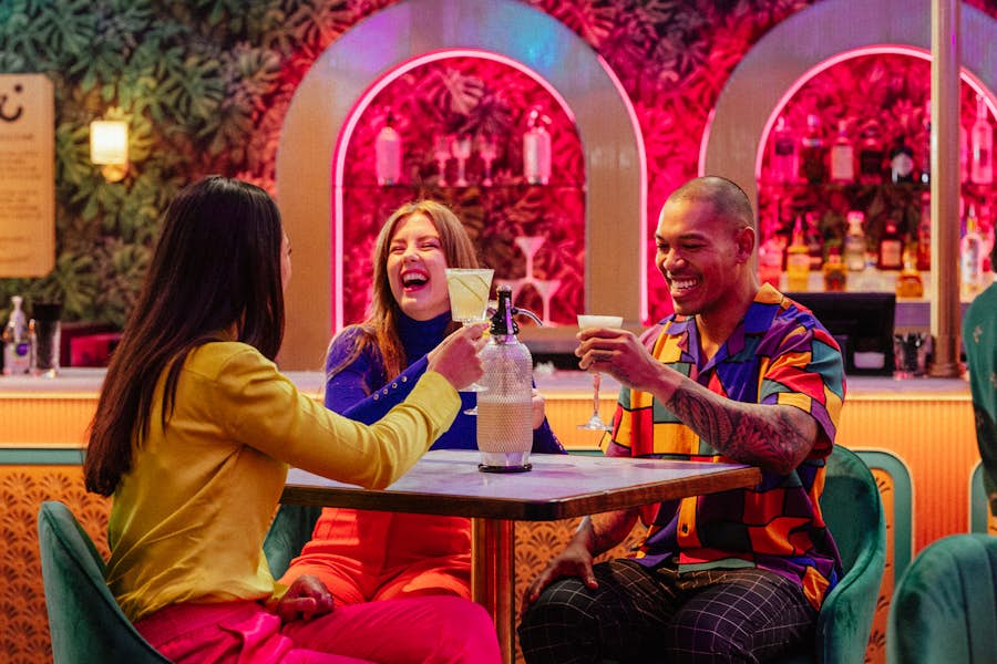 A group of three friends smiling and cheers-ing their drinks in the Hijinx Hotel lobby.