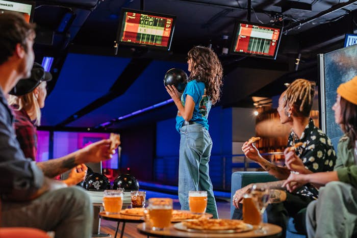 Woman preparing to bowl with group of friends looking on while eating pizza