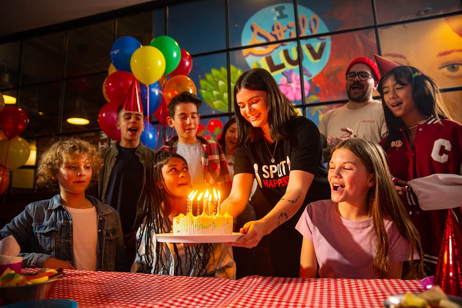 Strike party host lowering a child's birthday cake onto the table while friend and family sing