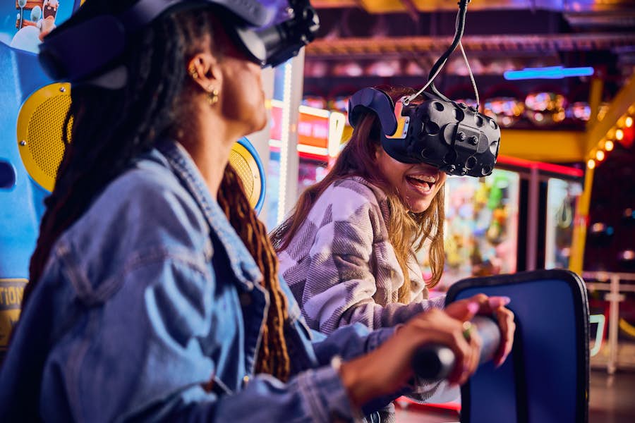 Two young women with VR headsets over their eyes smile and laugh while holding on to the game handle.