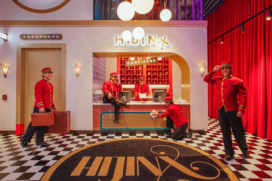 Image of the Hijinx Hotel check-in desk at Alexandria. The quirky concierge assistant can be seen in across the image 5 times, to the left of the image he is carrying luggage, next to that answering the phone while sitting on the desk, next, offering the viewer bespoke slippers, next cleaning the desk and on the far right tipping his red concierge  uniform cap.