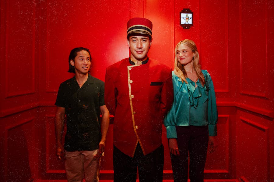 Hijinx Hotel in a concierge uniform stands between a male and female guest in the Hijinx Hotel 'elevator', smirking at the camera.