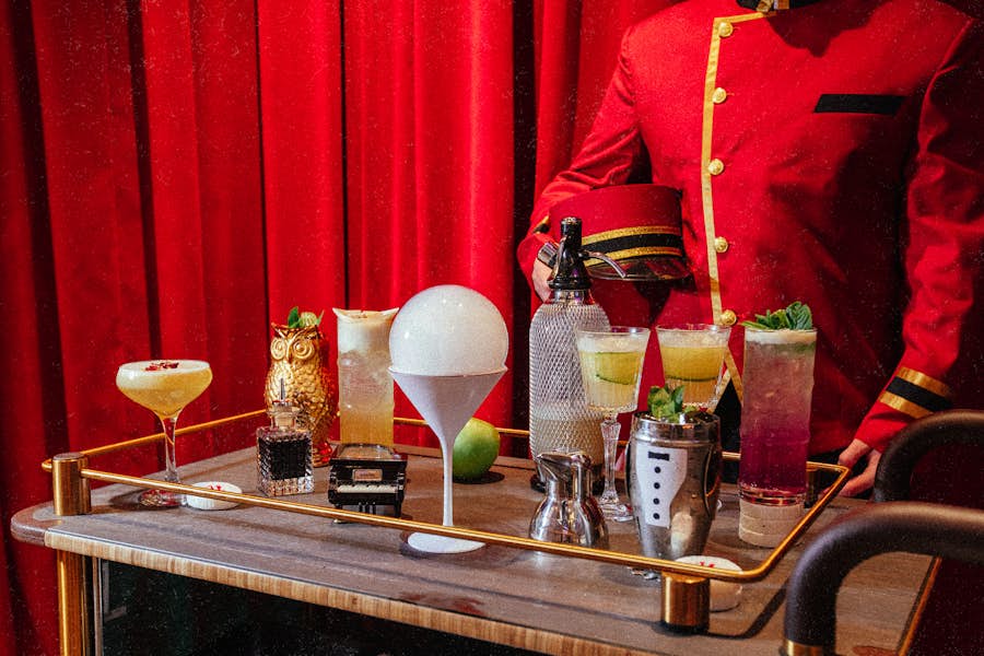 A bellhop in a red uniform stands behind a drinks cart with a selection of Hijinx Hotel signature cocktails.