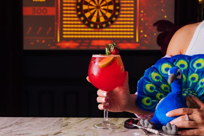 Woman holding a cocktail and plush peacock available at La Di Darts