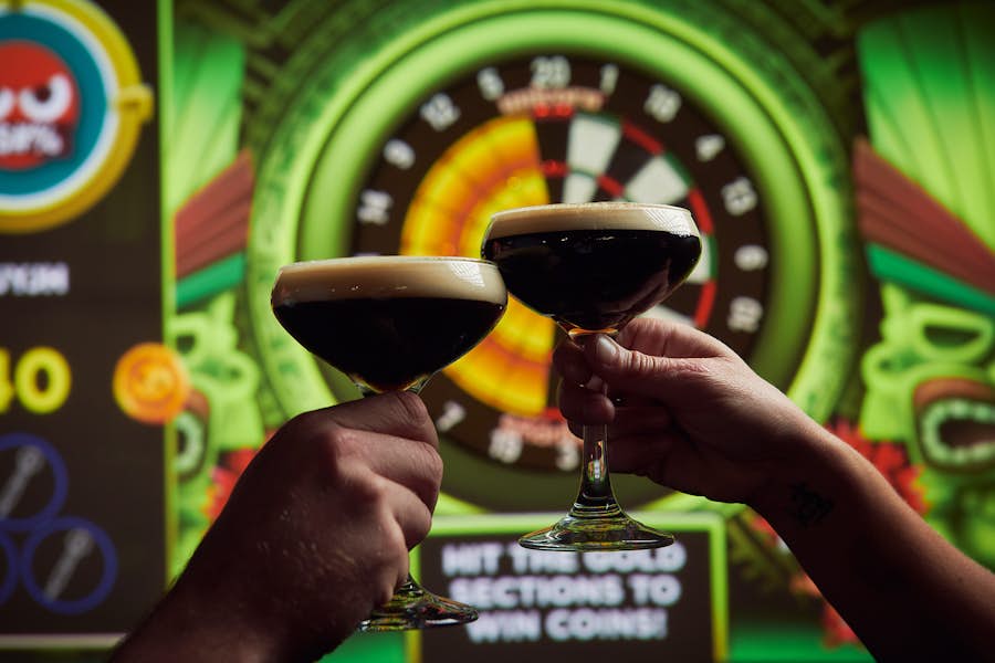 Two cocktail glasses being held in a toast with an interactive dart board in the background