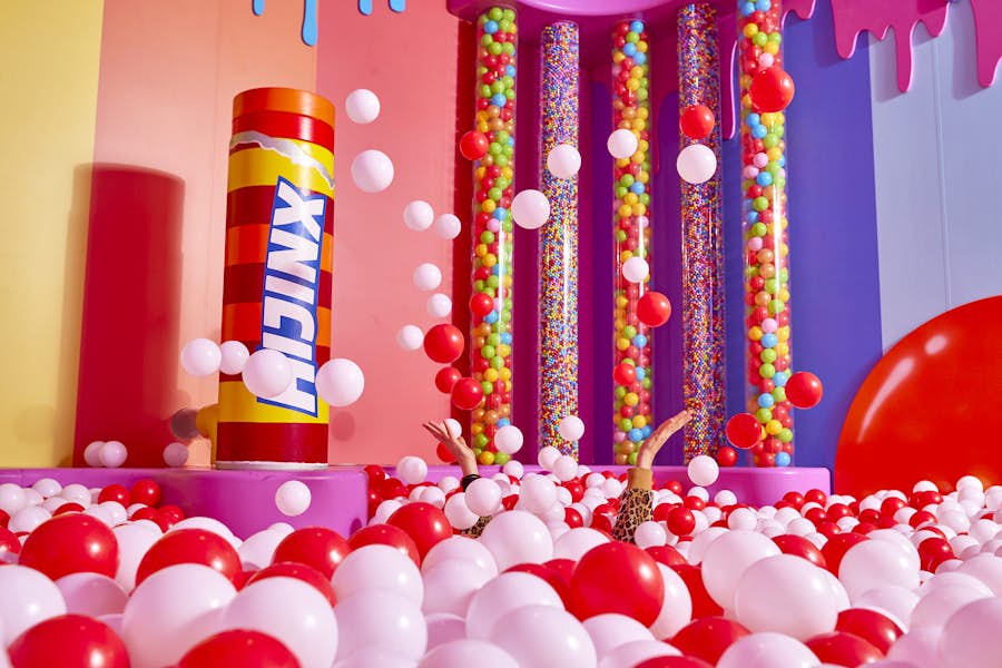 Candy themed ball pit challenge room at Hijinx Hotel with two hands popping out of the ball pit filled with red and white balls. The room is brightly coloured with pinks, purples, oranges and yellows colouring the walls.