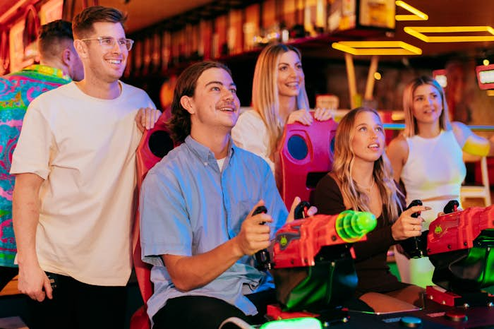 2 people playing Space Invaders arcade game with 3 people watching in the background