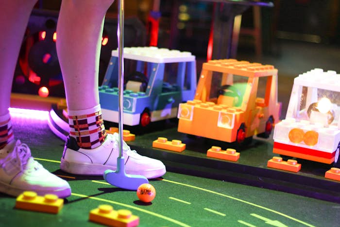 Person wearing white sneakers and long socks playing mini golf on a lego course with a blue putter and orange ball
