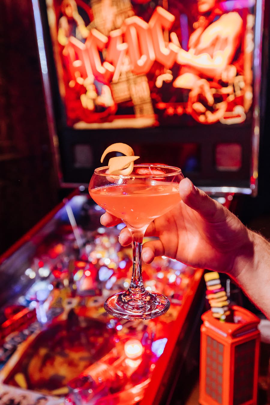Person holding a cocktail in the foreground with a pin-ball machine in the background