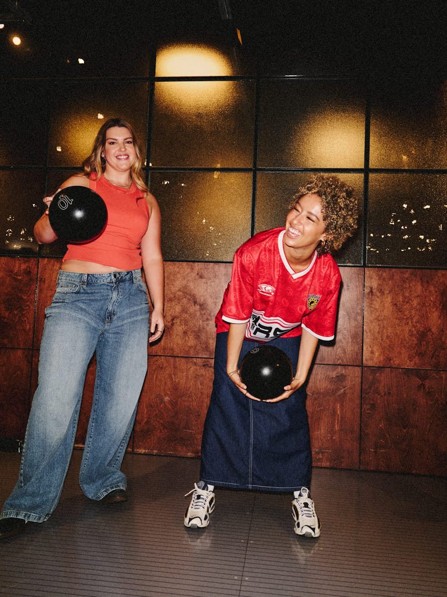 two girls in denim with bowling balls laughing and getting ready to bowl