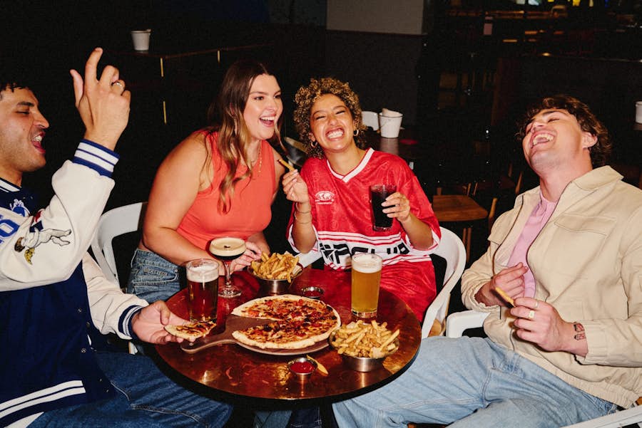 friends smiling and laughing while enjoying pizzas, fries, cocktails and beers