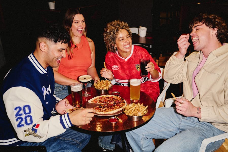 four friends at a table drinking beers and espresso martini cocktails while sharing pizza and bowls of fries