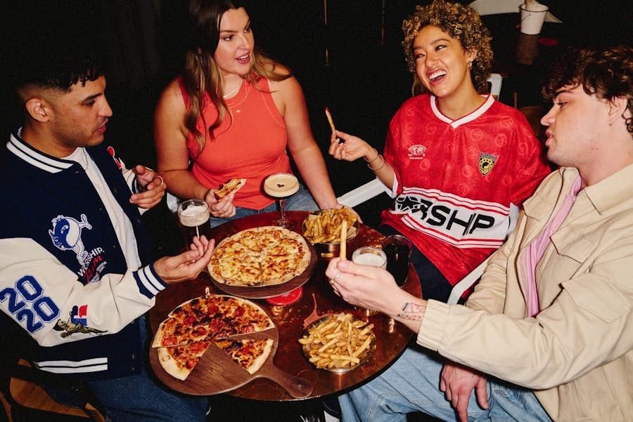 four friends at a table drinking beers and espresso martini cocktails while sharing pizza and bowls of fries