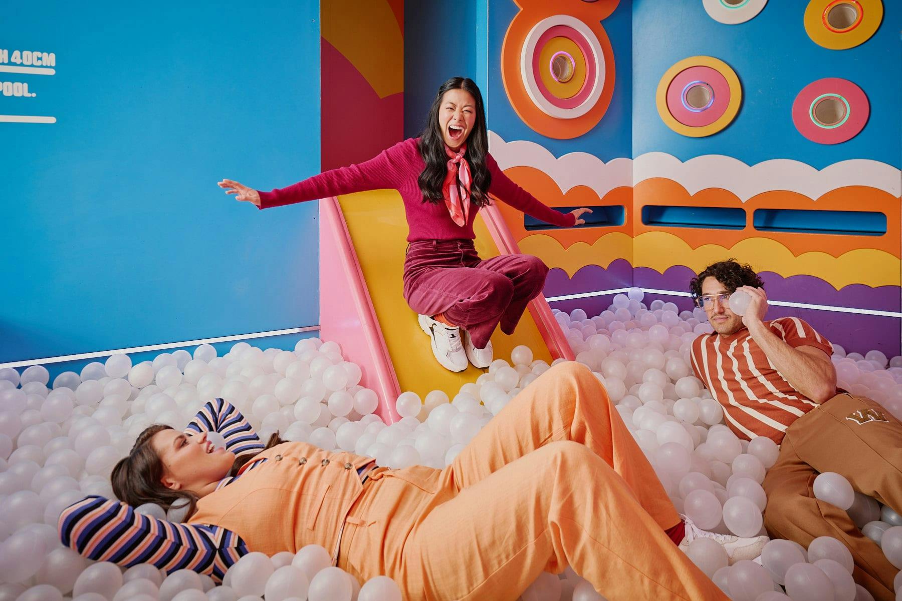 Hijinx Hotel Adore A Ball Pool room filled with white balls and multicoloured walls. There is a girl using the slide to get into the ball pit with her friends