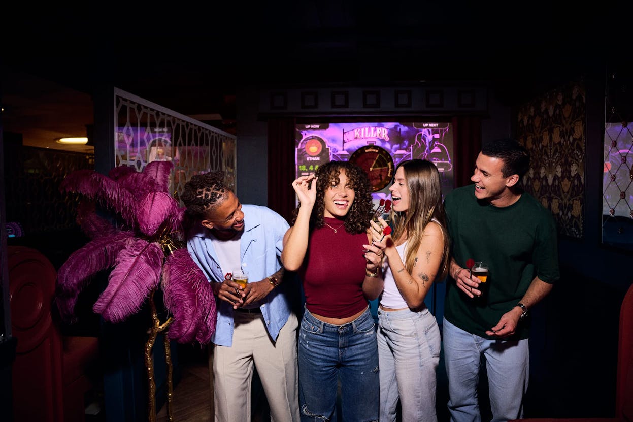 A group of four friends laughing, holding drinks and getting ready to throw darts towards the camera