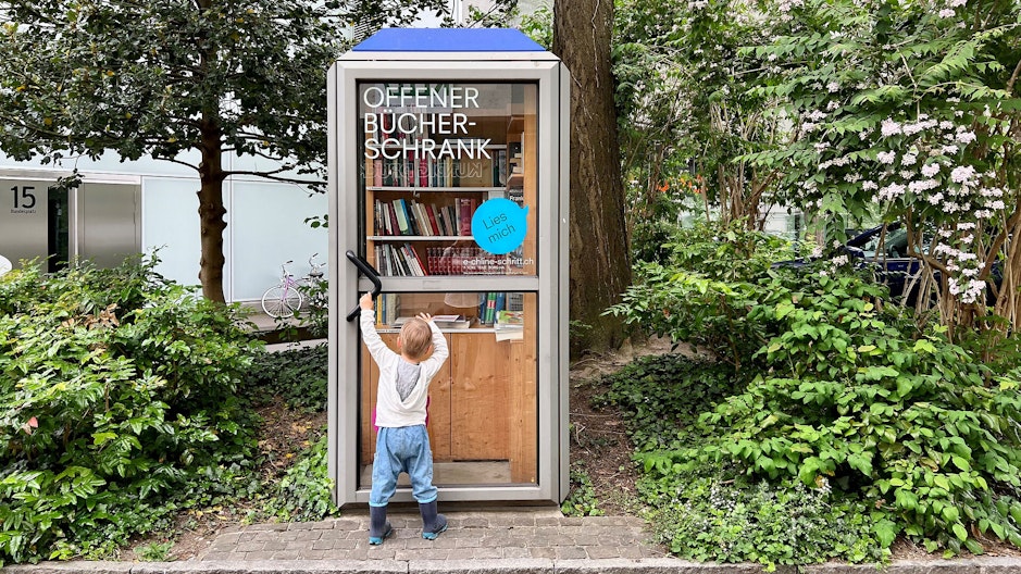 Ein Kind greift nach der Türe eines offenen Bücherschranks in einer ausgedienten Telefonzelle.