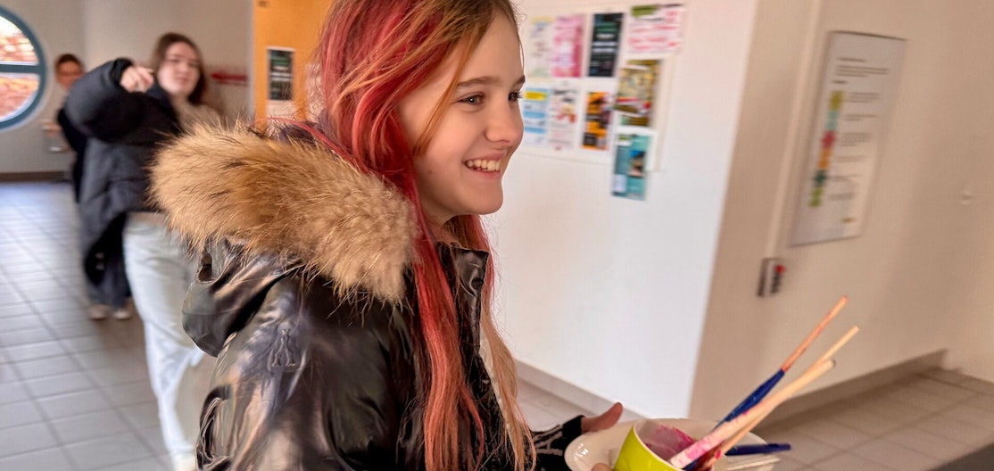 Schülerin mit Pinseln während der Projektwoche Ein Mädchen mit rot gefärbten Haaren und schwarzer Jacke mit Fellkragen lächelt, während es in einem gelben Becher mehrere bunte Pinsel trägt. Im Hintergrund sind andere Schüler:innen und ein Schulflur zu sehen.