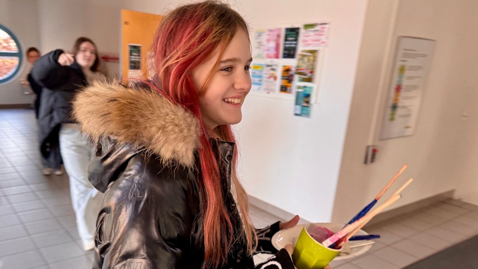 Ein Mädchen mit rot gefärbten Haaren und schwarzer Jacke mit Fellkragen lächelt, während es in einem gelben Becher mehrere bunte Pinsel trägt. Im Hintergrund sind andere Schüler:innen und ein Schulflur zu sehen.