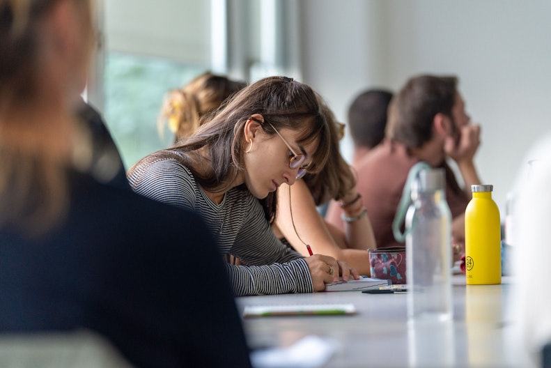 Eine junge Frau sitzt mit anderen Personen an einem Tisch und macht sich Notizen zur Weiterbildung.