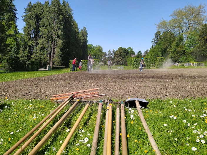 Auf einem Acker wird im Hintergrund eine Blumenwiese neu angesät. Im Vordergrund liegt Gartenwerkzeug.