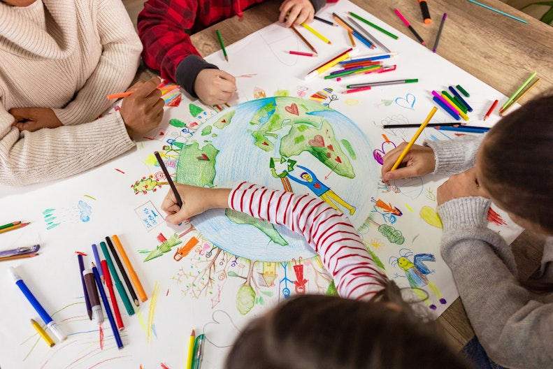 Kinderhände zeichnen zusammen ein Plakat, das die Welt mit vielen Figuren, Bäumen und Herzen zeigt