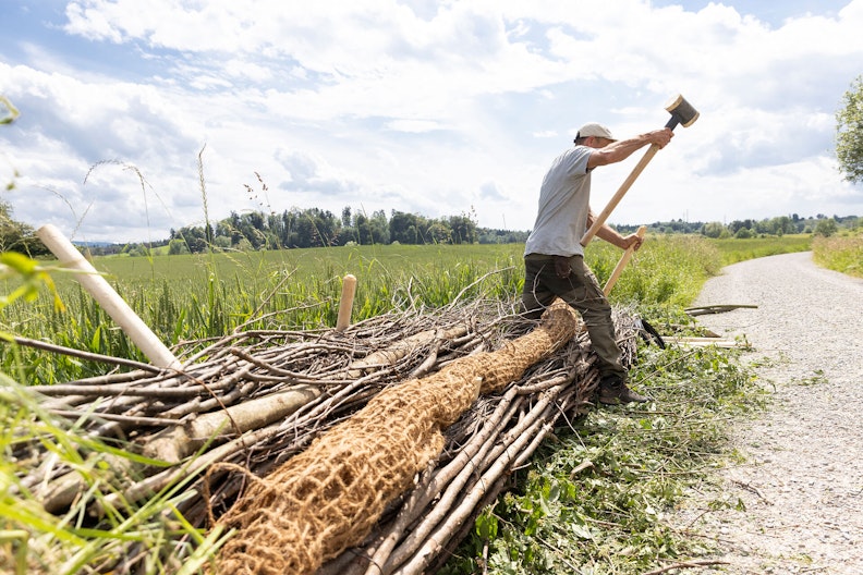 Ein angehender Gewässerwart steht breitbeinig über einem grossen Reisigbündel, das zu einer Faschine verbaut wird, und schlägt mit einem Hammer auf einen Pflock.