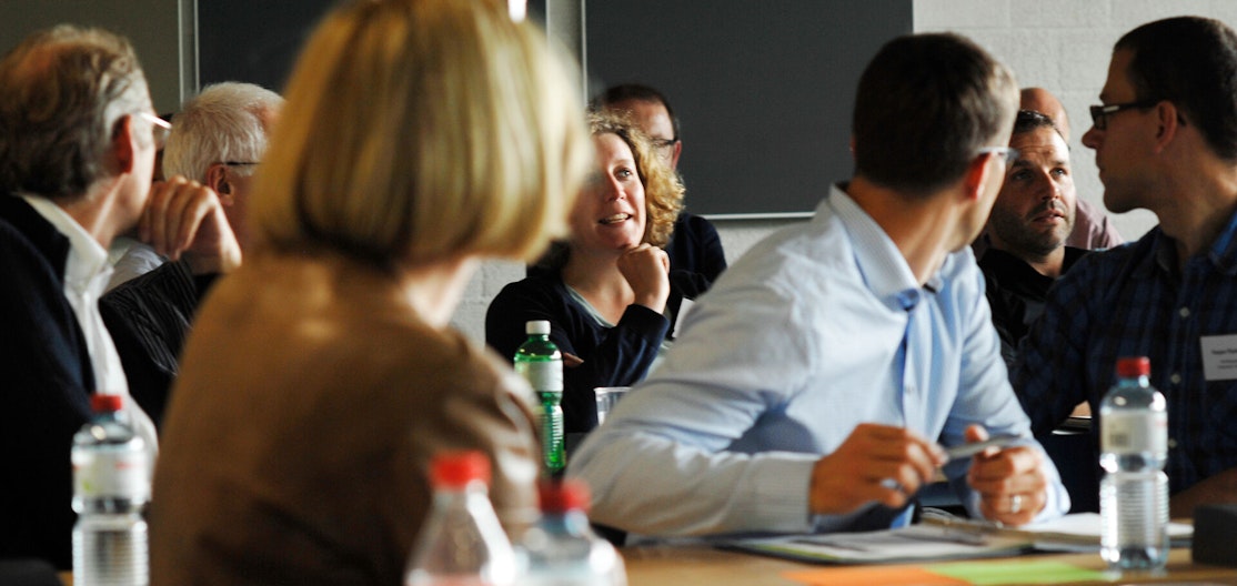 Achats publics responsables Un groupe de participants à un cours est assis à des tables dans une salle. Une femme assise dans une rangée au fond pose une question – tout le monde se tourne vers elle.
