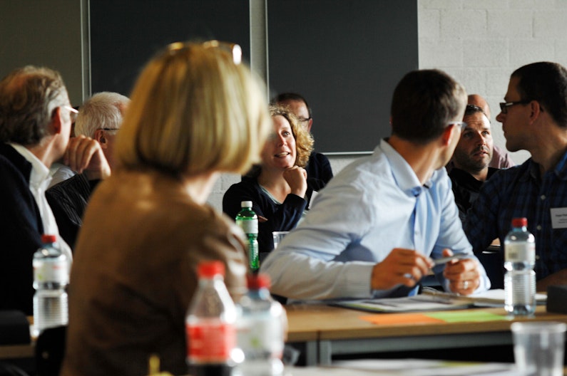 Eine Gruppe von Kursteilnehmern sitzt an Pulten in einem Raum. Eine Frau in einer hinteren Reihe stellt eine Frage – alle wenden sich ihr zu.