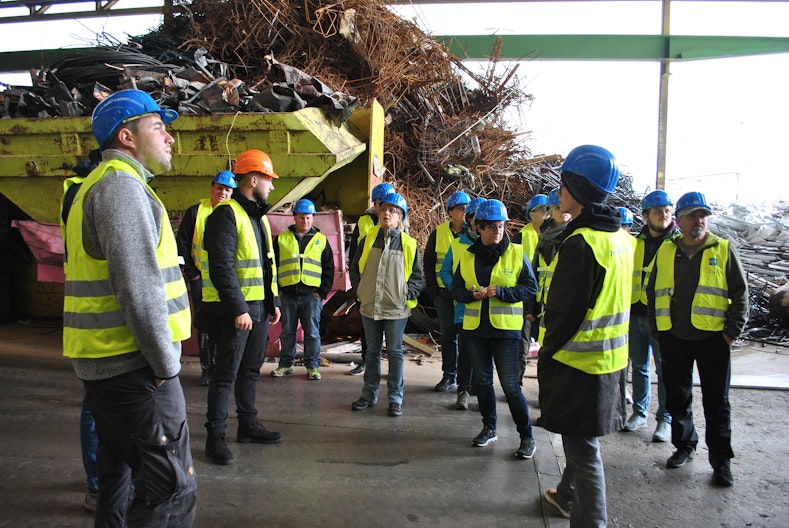 Kursteilnehmende mit gelber Warnweste und blauen Schutzhelfen stehen in einer Sammelstelle vor einem Abfallberg