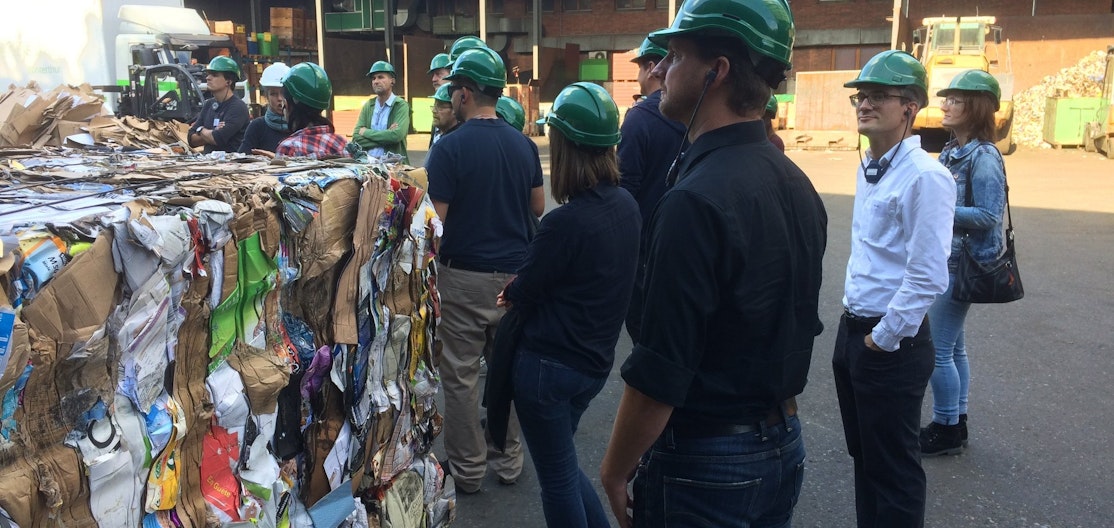 Weiterbildung zur Abfallbewirtschaftung Kursteilnehmende mit grünen Schutzhelmen stehen auf einer Sammelstelle neben einem Kubus aus gepresstem Karton.