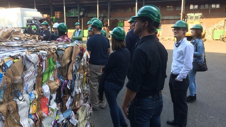 Kursteilnehmende mit grünen Schutzhelmen stehen auf einer Sammelstelle neben einem Kubus aus gepresstem Karton.