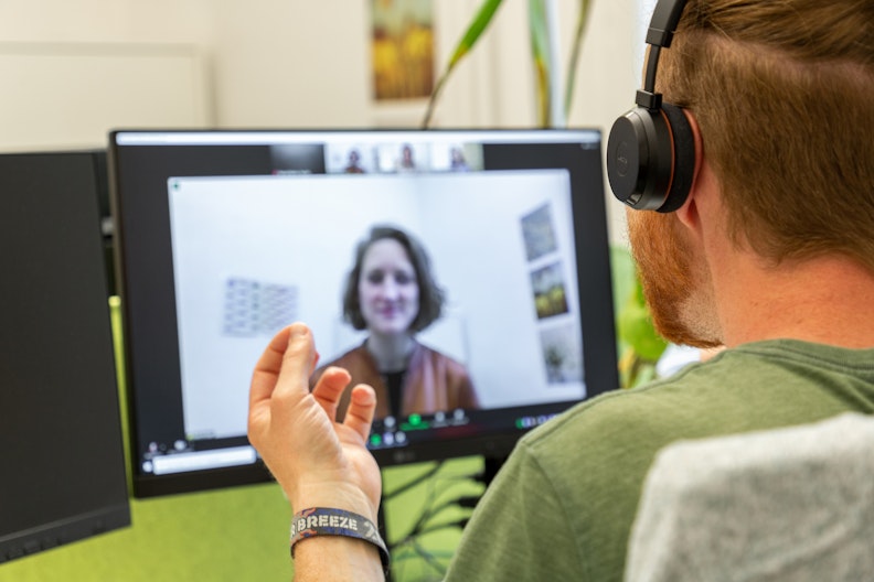 Ein Mann mit Headset sitzt vor dem Computer und führt eine Beratung durch. Auf dem Bildschirm ist unscharf eine Teilnehmerin zu erkennen.