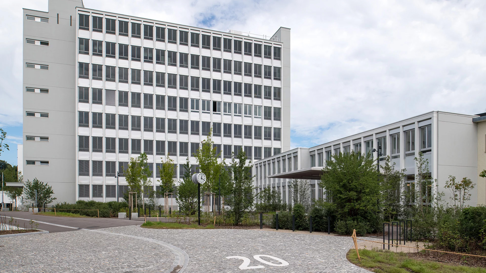 Im Hintergrund stehen zwei grosse Schulgebäude, davor ein offener Platz und dazwischen Grünflächen mit Bäumen und Sträuchern.