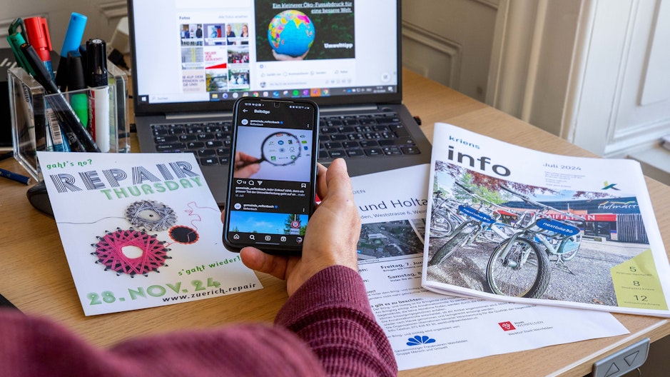 Eine Person hält ein Handy vor Laptop und Printmedien. Die 3 Geräte symbolisieren unterschiedliche Kommunikationskanäle für klimarelevante Informationen.